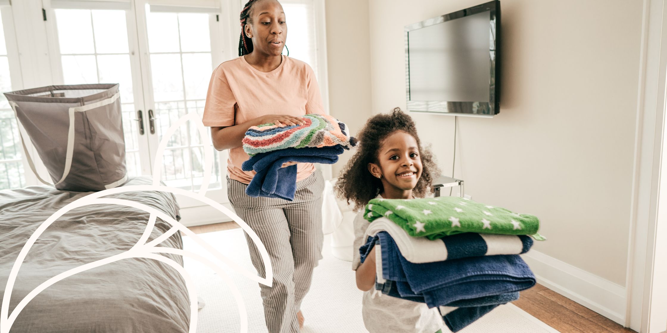 A mother and daughter in a bedroom, each holding a pile of clean, folded laundry from a basket on the bed, preparing to put it away together.