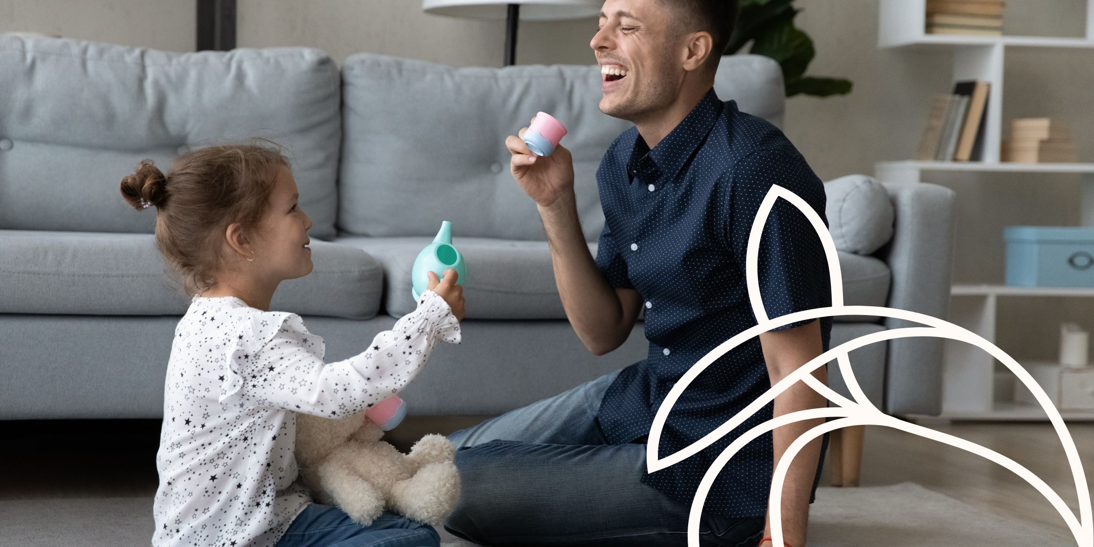 A dad sits on the living room floor across from his young daughter, laughing as he holds a toy tea cup. She grins brightly, holding a teddy bear in one hand and a play tea pot in the other, creating a heartwarming moment of connection and joy.