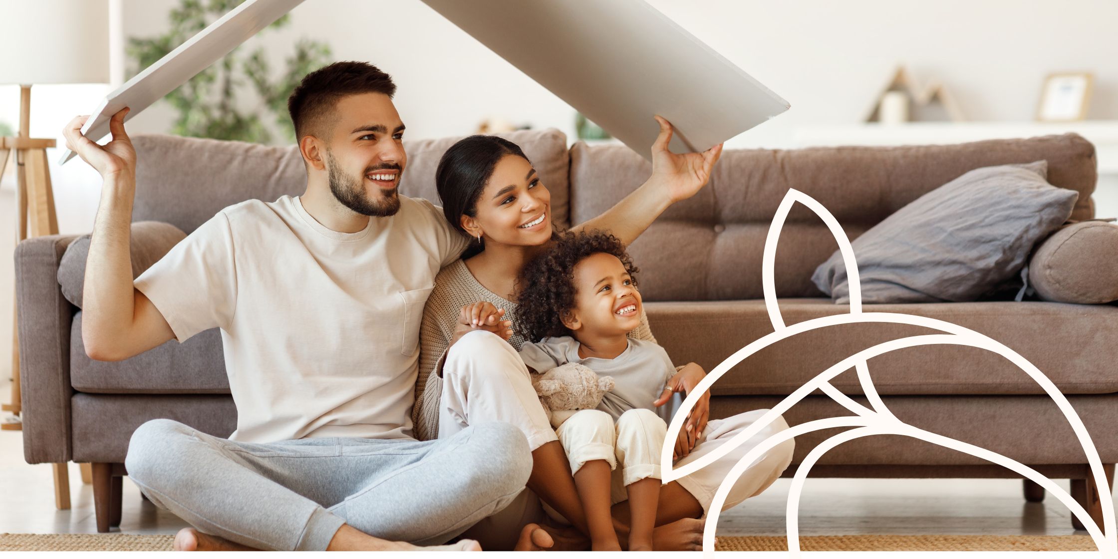 A family of three sits cross-legged on the living room floor in front of a couch. The father holds a large piece of cardboard, bent in half like a roofline, over the heads of the mother and daughter, symbolizing protection and togetherness in a safe home environment.
