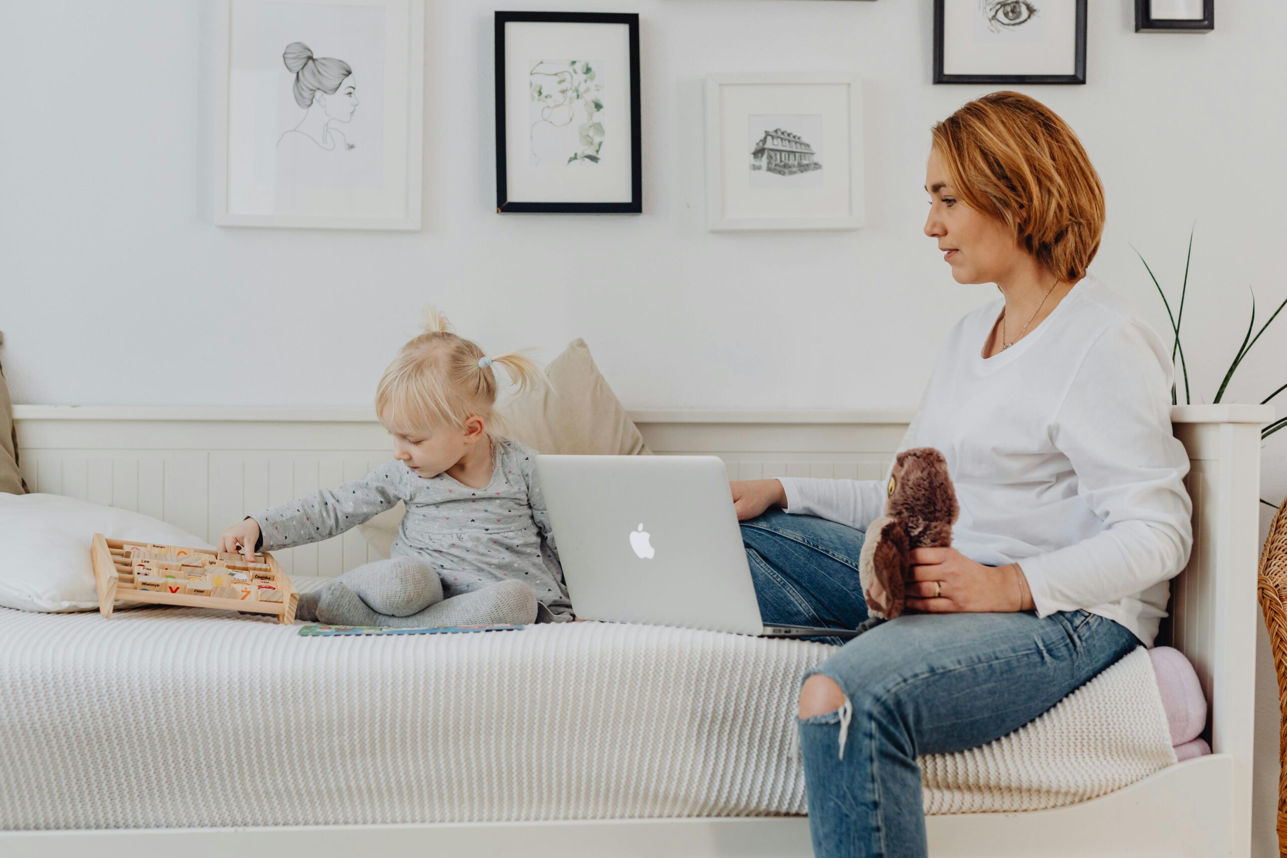 Pregnant mother working on a laptop while her toddler plays nearby on a daybed.