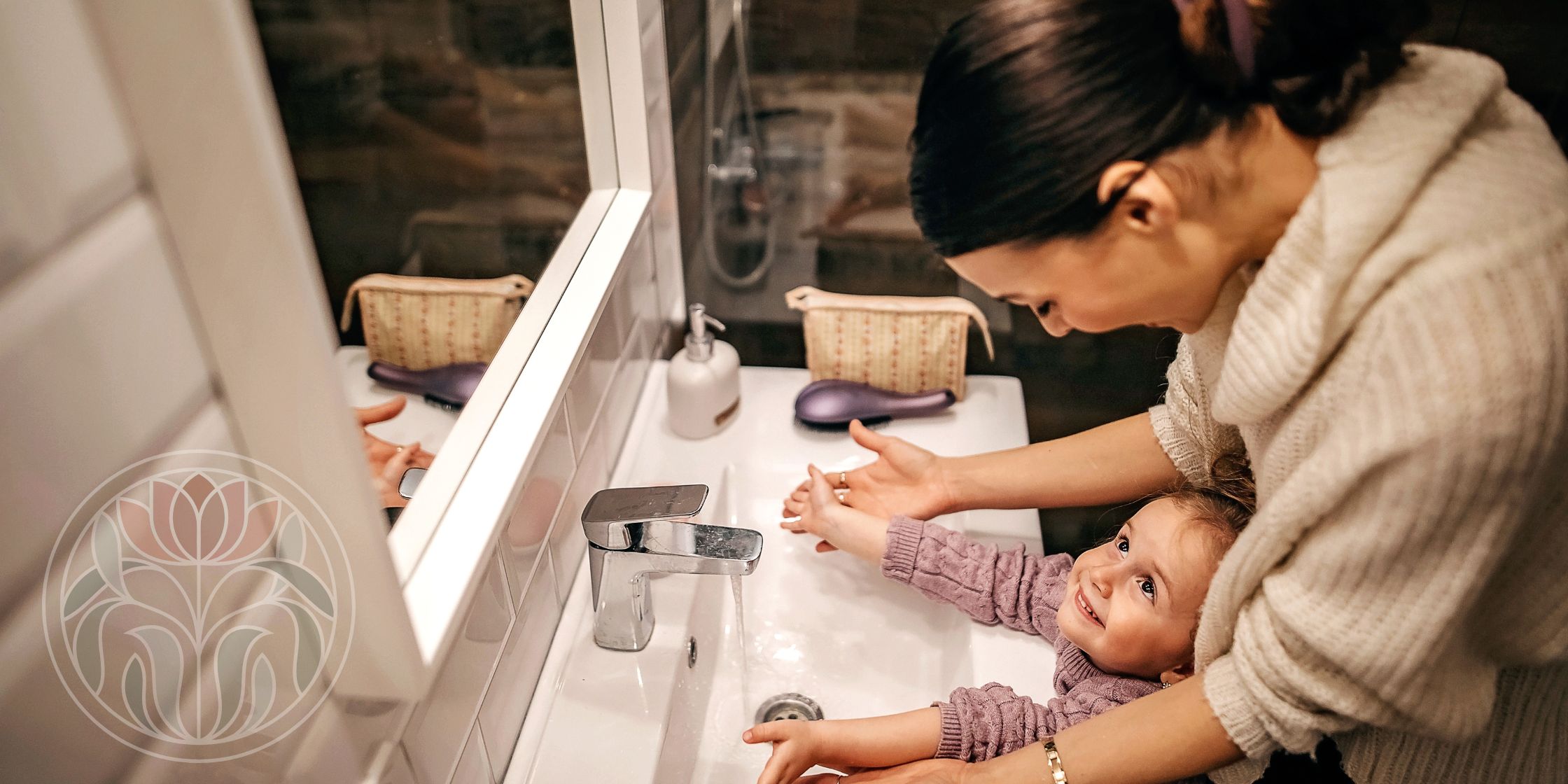 A mother stands behind her child at the bathroom sink, helping them get ready to wash their hands. The child looks up at the mother happily, capturing a warm morning moment.