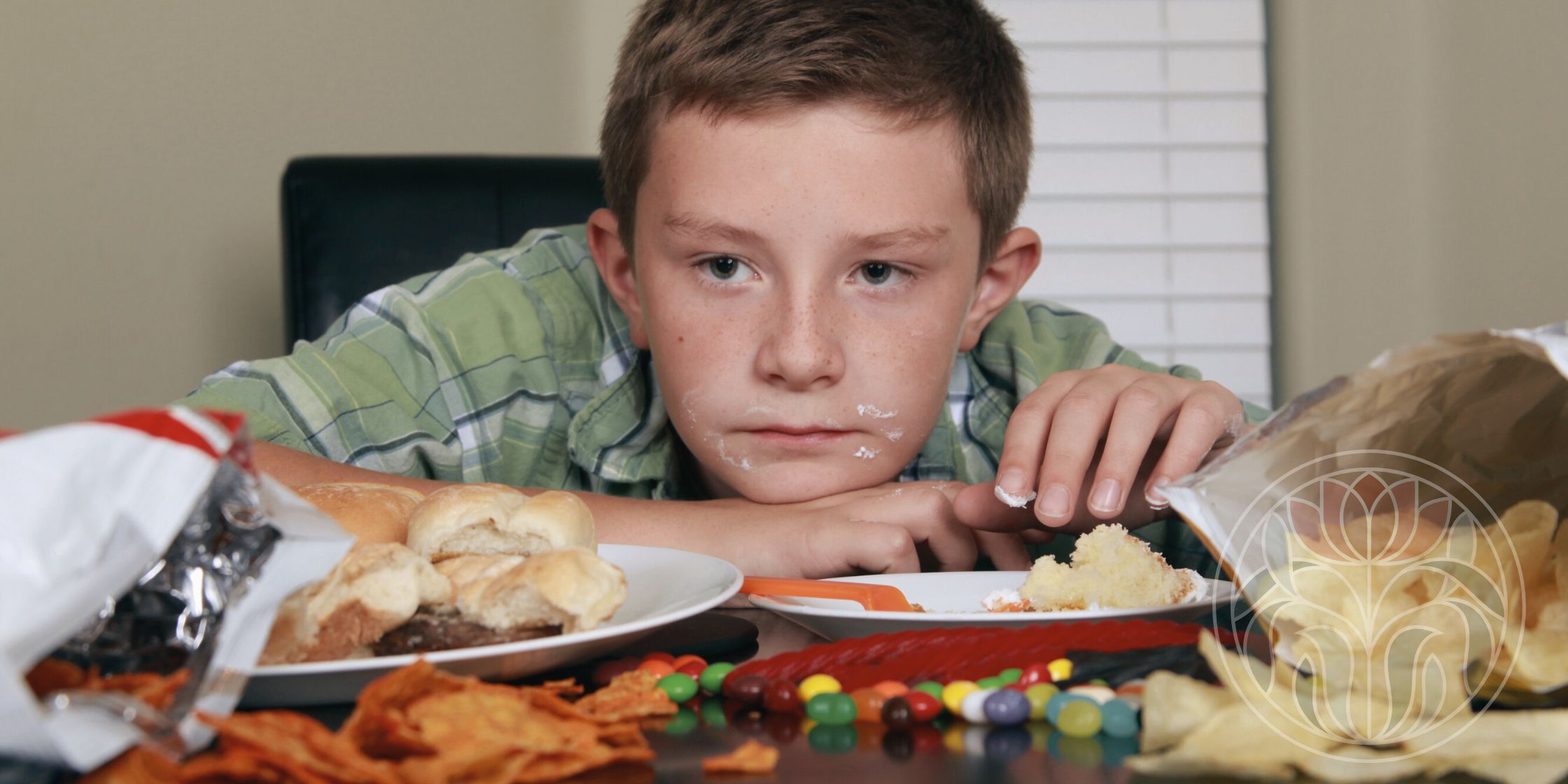 An adolescent boy sits at a table with a somber expression, a bit of frosting on his face. Before him are open bags of chips, spilled candies, an empty cupcake plate, burger sliders, and Twizzlers, visually representing the challenge of sensory-driven food choices.