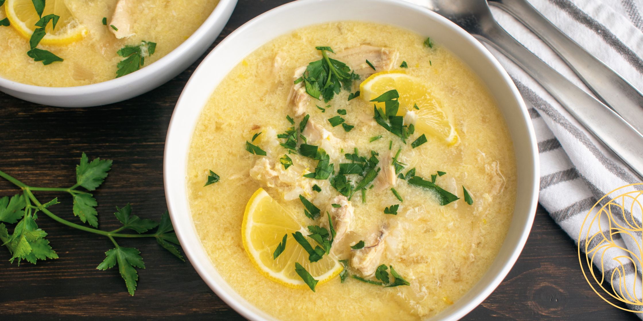 A white bowl of lemon chicken chowder topped with chopped parsley, featuring a tender chicken thigh in a golden broth with lemon slices on either side.