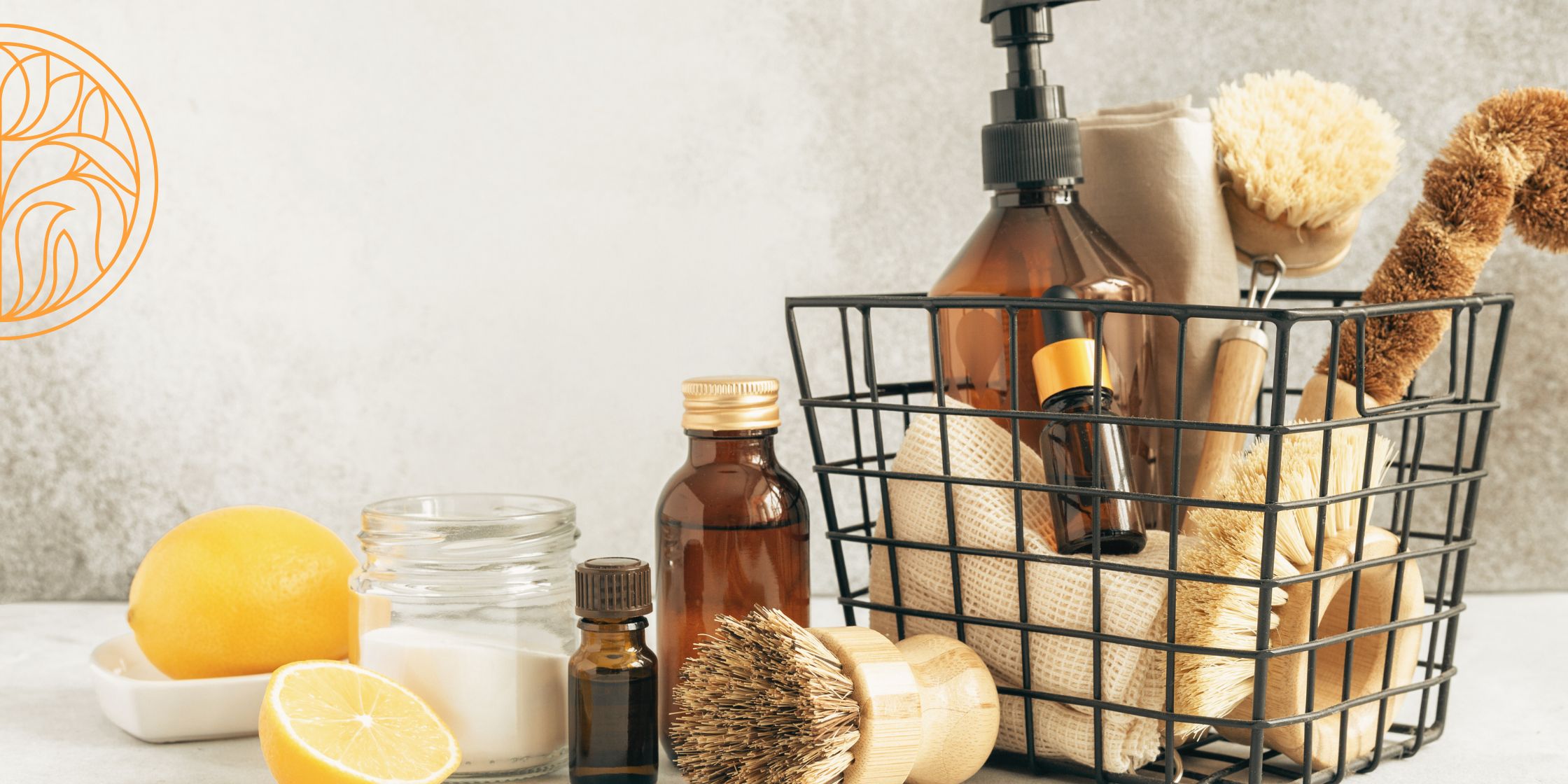 A wire basket filled with linen cleaning cloths, natural scrubbers with bamboo handles, and a brown glass spray bottle. Nearby, a cut lemon, a dish of cleaning powder, and another scrubber create a rustic and eco-friendly cleaning setup.